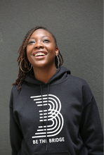 Woman wearing black hoodie with 'B' and 'Be The Bridge' in white lettering.
