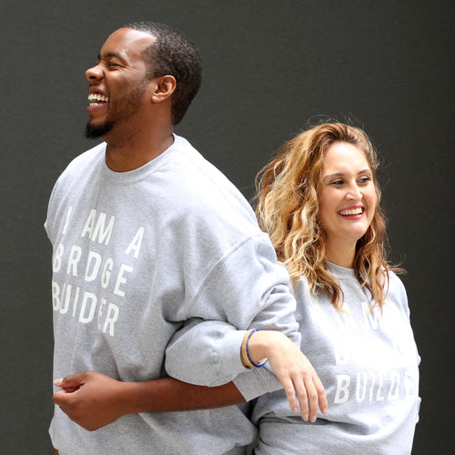Man and Woman linking arms wearing gray sweatshirt with 'I Am A Bridge Builder' in white lettering.